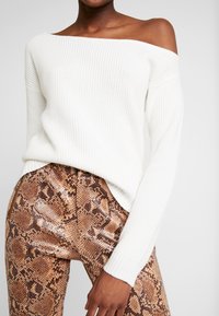 Kvinna iklädd en off-shoulder vit ribbad tröja och högmidjade bruna byxor med ormtryck och dragkedjedetaljer.