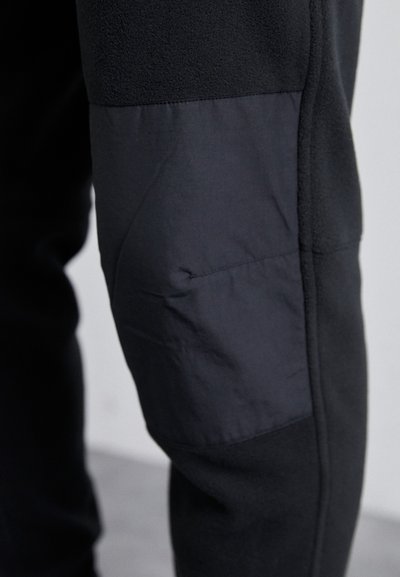 Calças de jogging pretas com uma combinação de materiais pretos lisos e texturizados. Apresenta um remendo no joelho com um ligeiro brilho e detalhes cosidos.