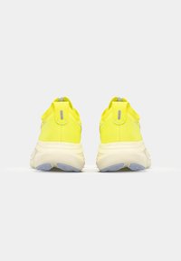 Gelbe Sportschuhe mit einer strukturierten Oberseite, Ziehlappen an der Ferse und einem gepolsterten Kragen. Dicke Sohle mit einem grau-blauen Akzent an der Basis.