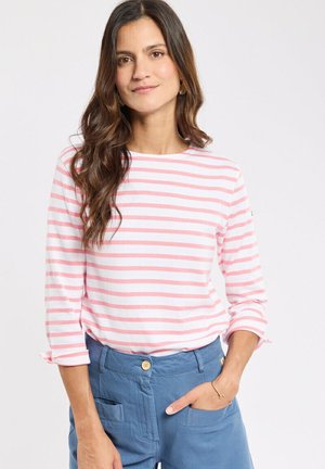 Femme aux longs cheveux bruns portant un haut à manches longues rayé rose et blanc et un pantalon bleu, debout avec une main dans la poche.