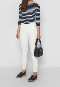 Mujer con una blusa de rayas en azul marino y blanco de manga larga, jeans blancos, mocasines azul marino y sosteniendo un bolso azul marino con interior azul claro.