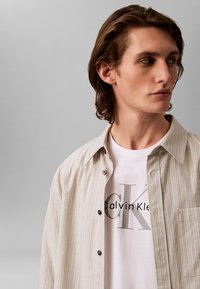 Lys beige, stripete skjorte med knappelukking over en hvit T-skjorte med grå "Calvin Klein"-logo. Glatt tekstur, avslappet passform.