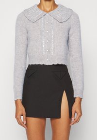 Ljusgrå stickad kort cardigan med en vågig nederkant, dekorativa knappar och en strukturerad krage, kombinerad med en svart minikjol med snedställda fickor.