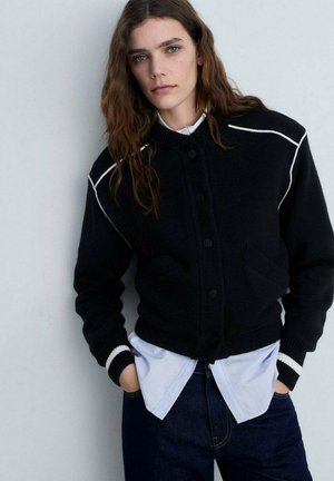 Schwarze Strickjacke mit weißen Akzenten, mit einer Knopfleiste vorne, zwei Fronttaschen und gerippten Bündchen, übereinander getragen mit einem hellblauen Hemd.