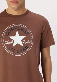 Barna, pamutból készült póló, kerek nyakkivágással, amelyen egy kör alakú fehér logó található, csillaggal és fekete szöveggel: "CONVERSE ALL STAR".