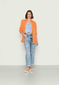Blazer laranja sobre um top de flores azul claro, combinado com jeans claros desgastados e sandálias brancas. Design simples, look casual.