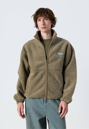 Junge Person mit kurzen braunen Haaren, die eine hellbraune Fleecejacke mit Reißverschluss und blaue Jeans vor einem einfarbigen Hintergrund trägt.