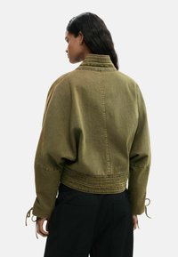 Femme aux longs cheveux bruns portant une veste courte vert olive et un pantalon noir, dos à la caméra sur un fond uni.