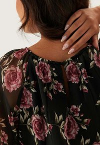 Svart blommig blus med en volangkrage, rosa rosor och gröna blad. Baksidan har en nyckelhålsdetalj med knappstängning.