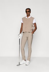 Pulover bej fără mâneci, tricotat cu model în cablu, peste o cămașă polo albă, asortat cu pantaloni cremuți conici și pantofi albi de golf, ținând un club de golf.