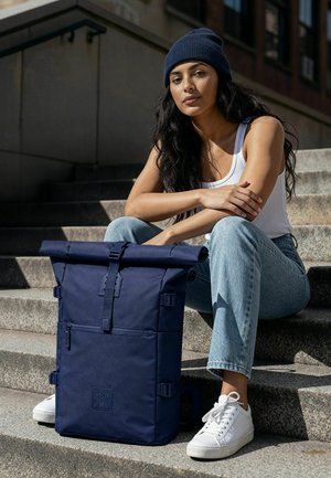 Junge Frau mit einer marineblauen Mütze, einem weißen Tanktop und Jeans sitzt auf Außentreppen, neben ihr ein großer marineblauer Rolltop-Rucksack.