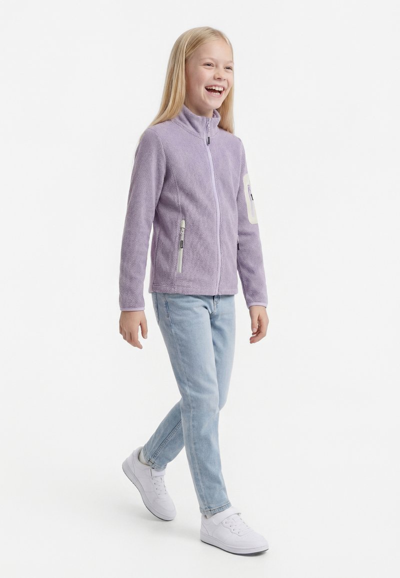 Enfant souriant marchant, portant une veste zippée violet clair, un jean bleu clair et des baskets blanches sur un fond blanc uni.
