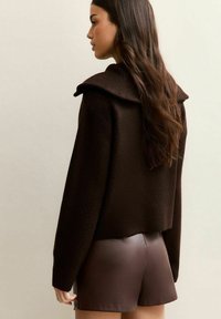 Brązowy sweter z fakturowanej dzianiny z oversize'owym kołnierzem, zestawiony z gładkimi brązowymi skórzanymi szortami, widok z tyłu.