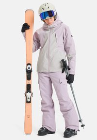 L'ensemble de vêtements de ski comprend une veste violet clair avec des accents gris et un pantalon assorti. Le skieur tient un ski orange et des bâtons gris, portant des lunettes et un casque.