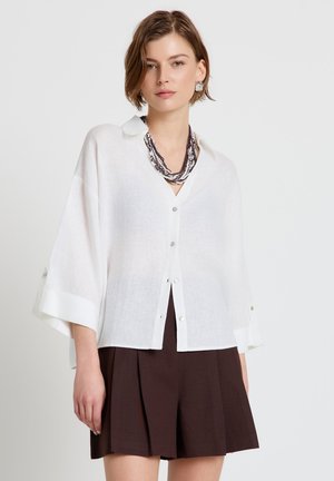 Frau trägt locker sitzendes weißes Hemd mit weiten Ärmeln, dunkle plissierte Shorts und mehrlagige Perlenkette vor schlichtem Hintergrund.