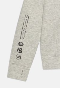 Grauer Pullover mit weicher Textur. Enthält schwarze Druckgrafiken auf dem Ärmel, darunter ein Logo, ein Tier und die Nummer 92.