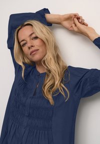 Camicia button-up blu navy con dettagli arricciati sul petto e maniche a sbuffo. Tessuto morbido, vestibilità comoda e scollo a chiave.