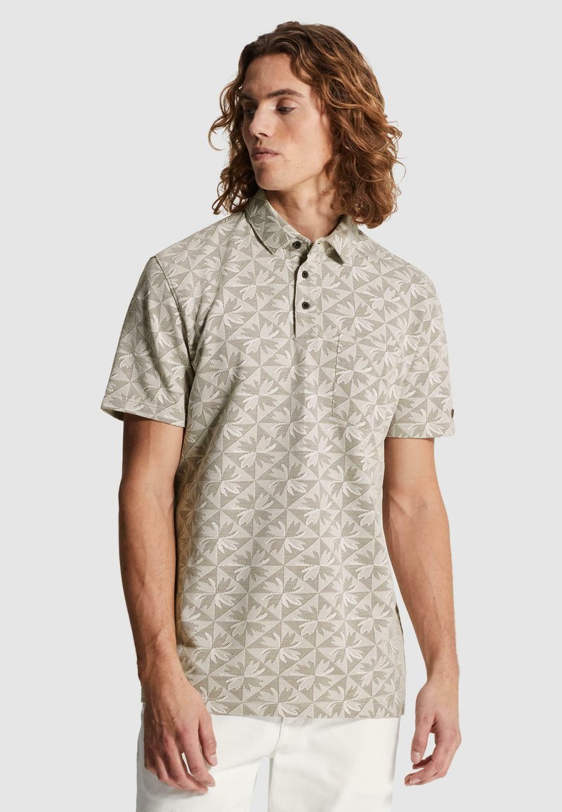 Homme aux cheveux bouclés mi-longs, portant un polo beige à manches courtes avec un motif floral géométrique et un pantalon blanc.