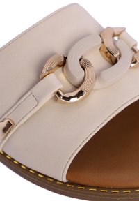 Sandalia deslizante de cuero beige que presenta una tira ancha con un acento de cadena dorada y texturizada blanca. Plantilla marrón con detalle de costura amarilla.