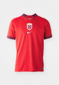 Maglia Nike rossa con colletto circolare, maniche corte e rifiniture decorative blu. Logo della squadra centrato con un design a scudo e testo.