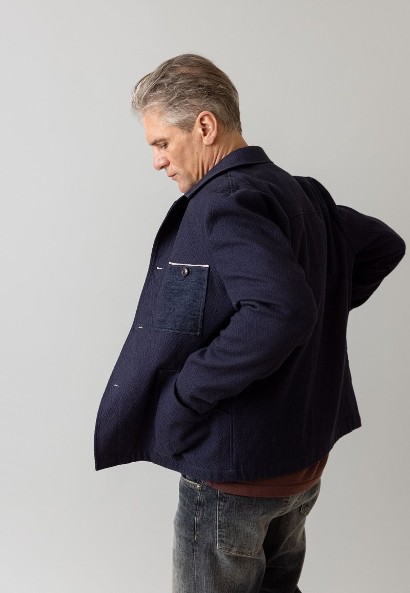 Navy blue jacket with textured fabric, featuring a denim pocket detail. The design includes a collar and hidden closure buttons.