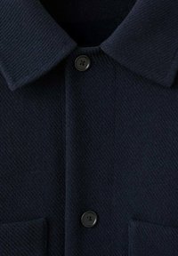 Temno mornarsko modra volna s srajco s ovratnikom, teksturirano tkanino, en žep na prsih in črnim gumbom na ovratniku.