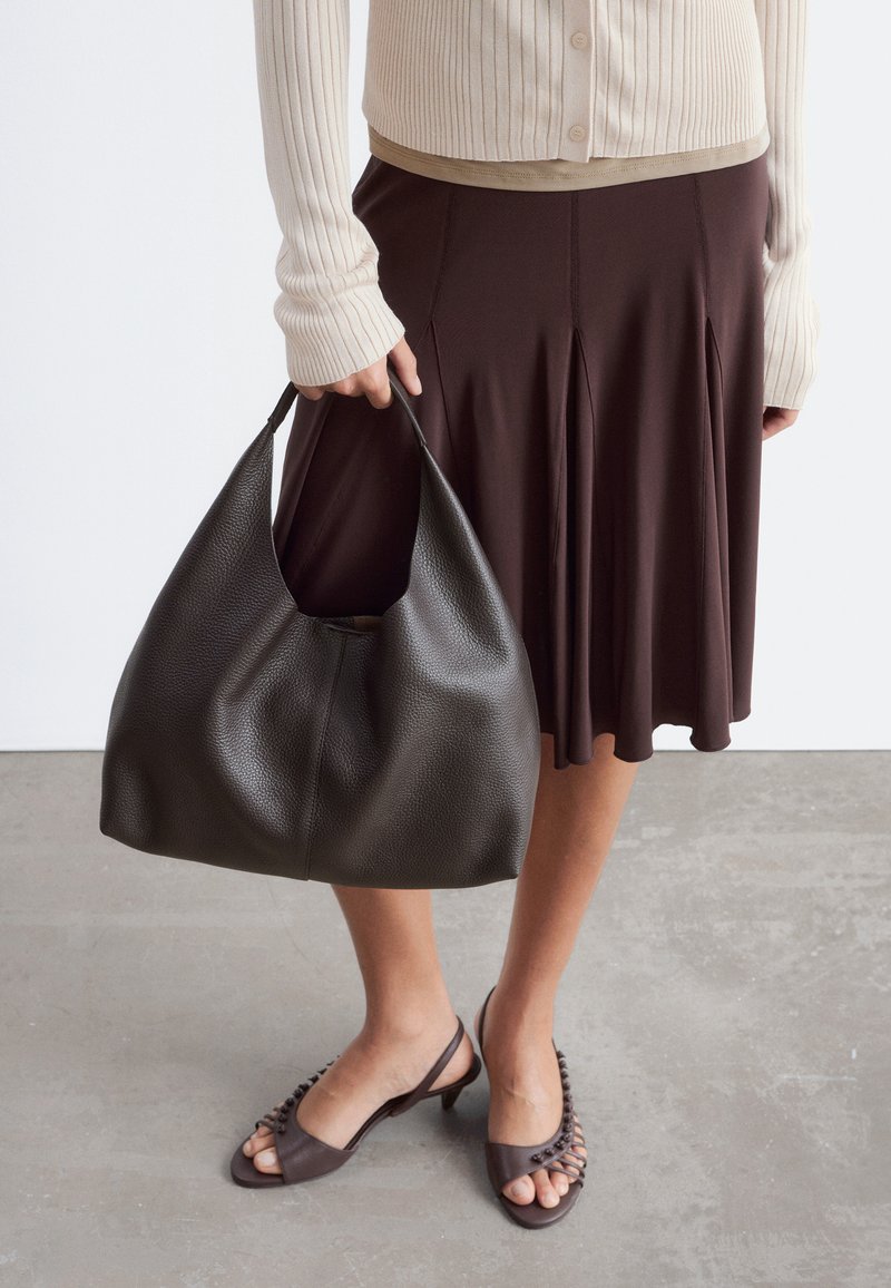 Personne portant un cardigan beige en maille côtelée, une jupe marron et des sandales à talons, tient à ses côtés un grand sac à main noir en cuir texturé.