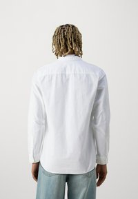 Camicia bianca a maniche lunghe con colletto, caratterizzata da un orlo dritto, polsini con bottoni e una texture liscia, indossata con jeans blu chiaro.