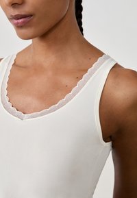 Primo piano di una donna che indossa una canotta color crema con bordi in pizzo scallop lungo il colletto e le spalline.