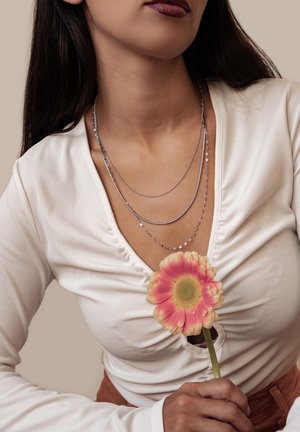 Frau mit dunklem Haar trägt geschichtete silberne Halsketten und ein weißes, gerafftes Oberteil, hält eine rosa Gerbera-Blume nahe ihrer Brust.