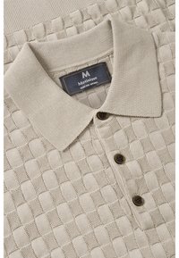 Beige adīts polo krekls ar teksturētu kvadrātu rakstu, klasisku apkakli un trim brūniem pogām pie apakšmalas.