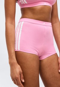 Γυναίκα που φοράει ροζ αθλητικές σορτς και ασορτί αθλητικό σουτιέν με λευκές ρίγες, στέκεται μπροστά σε απλό φωτεινό φόντο.