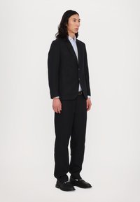 Schwarzer Anzug mit taillierter Passform, ausgestattet mit eingeschlagenen Revers, zwei vorderen Taschen und einem hellblauen Hemd darunter. Schwarze Schuhe runden den Look ab.
