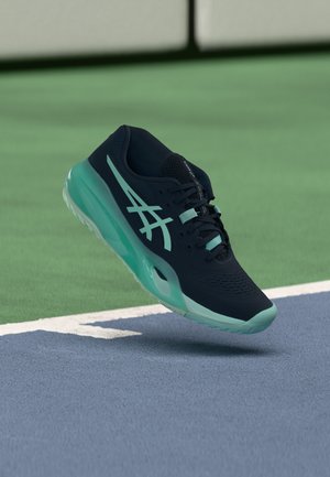 Dark blue and light blue athletic shoe hovering above a tennis court surface near the white boundary line.