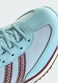 Hellblauer Sneaker mit weißen Mesh-Paneelen, hellblauen Schnürsenkeln und drei zickzack-gesteppten kastanienbraunen Streifen an der Seite.