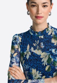 Modra cvetna obleka z nabrano teksturo, volančnim izrezom in dolgimi rokavi. Cvetni vzorci vključujejo bele in zelene poudarke. Srebrni uhani.