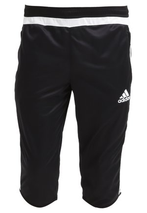 Mężczyzna w białej koszulce Adidas, czarnych krótkich spodniach Adidas, białych skarpetkach Adidas i białych korkach, stoi na jednolitym tle.