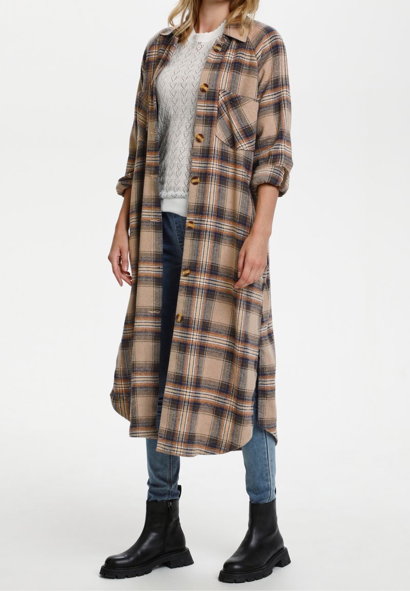 Lång skjortjacka i beige, brun och marinblå, med knäppning, bröstficka och uppkavlade ärmar, kombinerad med jeans och svarta stövlar.