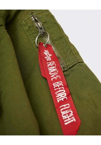 La tela de la chaqueta de color verde oliva presenta una cremallera de metal y una etiqueta roja de "RETIRAR ANTES DEL VUELO" con texto blanco, unida mediante un anillo plateado.