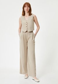Gilet sans manches beige avec des boutons, associé à un pantalon large. Texture lisse, couleur neutre et deux poches avant.