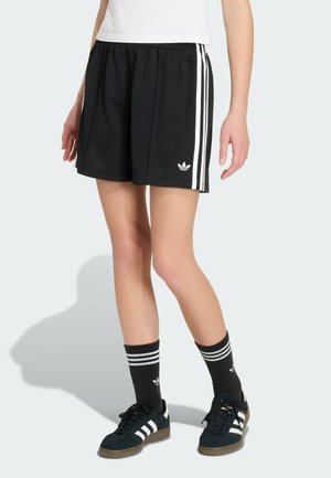 Henkilö, jolla on päällään mustat Adidas-shortsit valkoisilla raidoilla, mustat Adidas-sukat raitoineen ja mustat Adidas-tennarit valkoisilla raidoilla.