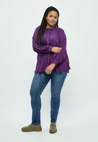Blusa lunga viola a maniche lunghe con dettagli plissettati, vestibilità comoda e chiusura con bottoni, abbinata a jeans slim fit e scarpe chunky beige.