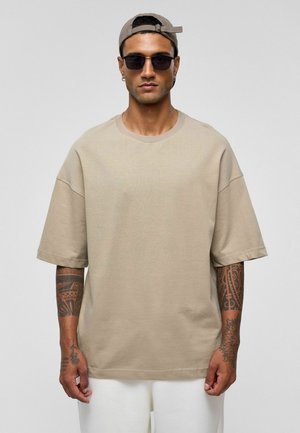 Lichtbeige oversized T-shirt met korte mouwen, ronde hals en een ontspannen pasvorm. Gladde stoftextuur, gedragen met witte broeken en een zonnebril.
