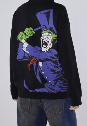 Sweatshirt noir arborant un graphique d'un personnage de dessin animé avec des cheveux verts, riant et tenant un haut-de-forme, avec des accents violets et rouges.