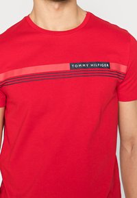 Rød bomulds T-shirt med korte ærmer, rund hals, med et centralt sort og rødt bånd mærket "TOMMY HILFIGER" og vandrette striber.