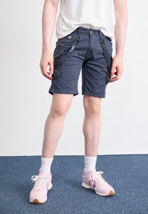 Marine cargoshorts met zijzakken, knoopsluiting en riemlussen. Draag met roze sneakers en witte crew-sokken op een blauw tapijt.