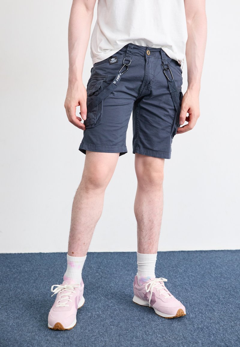 Marine cargo shorts med sidelommer, knaplukning og bæltestropper. Båret med lyserøde sneakers og hvide ankelstrømper på et blåt tæppe.