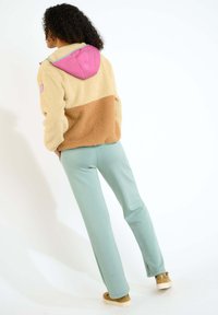 Giacca in fleece color tan e crema con cappuccio rosa, abbinata a pantaloni verde chiaro. La giacca presenta una superficie testurizzata e una decorazione con patch.