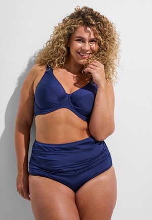 Kobieta o kręconych włosach uśmiechająca się, w granatowym dwuczęściowym stroju kąpielowym, stojąca przy białej ścianie z ręką blisko twarzy.