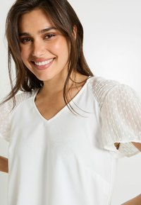 Top blanco con escote en V y mangas cortas abullonadas, hecho de una tela transparente y texturizada con un patrón de puntos. Acabado general suave.
